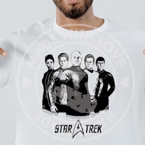 Puede incluir: Camiseta blanca con un gráfico en blanco y negro de cinco personajes de Star Trek. El logotipo de Star Trek está debajo de la imagen. Una persona señala la camiseta.