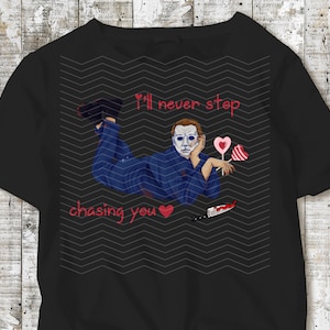 May include: Black t-shirt featuring a graphic of a character in a blue jumpsuit and a white mask, with the text "I'll never stop chasing you." The design includes heart-shaped lollipops and a bloody knife.