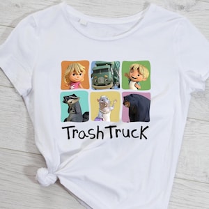 Puede incluir: Camiseta blanca con un gráfico colorido de personajes de la serie animada "Trash Truck". El diseño incluye seis imágenes de dibujos animados de una niña, un camión, un niño, un mapache, una rata y un oso, con las palabras "Trash Truck" en negro.