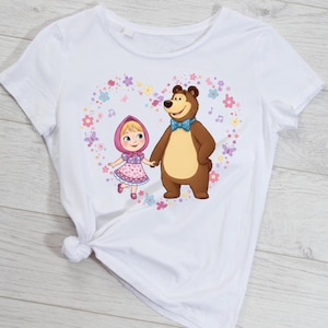 May include: White t-shirt featuring a cartoon illustration of a girl in a pink dress and a brown bear holding hands inside a heart-shaped floral design. The bear wears a blue bow tie. The design includes musical notes and butterflies.