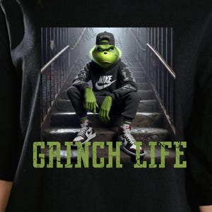 Peut inclure: Un personnage de Grinch vert portant un sweat-shirt noir avec un logo Nike blanc et des baskets noires et blanches. Le Grinch est assis sur un escalier avec le texte "Grinch Life" en lettres vertes.