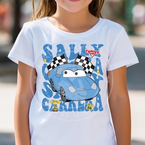 May include: White t-shirt featuring a blue cartoon car with checkered flags, the text "SALLY CARERRA", and the Cars logo. The design includes a Route 66 sign and a trophy. The shirt is likely for children.
