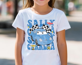 May include: White t-shirt featuring a blue cartoon car with checkered flags, the text "SALLY CARERRA", and the Cars logo. The design includes a Route 66 sign and a trophy. The shirt is likely for children.