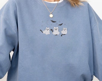 May include: A light blue sweatshirt with a graphic of three white cats dressed as ghosts with a crescent moon and bats in the background. The text "Blue Jean" is in the top right corner of the image.