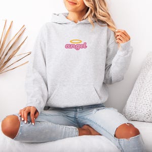 May include: A light gray hoodie with a front pocket and a hood. The word "angel" is embroidered on the chest in pink with a gold halo above it. The model is wearing light blue jeans with ripped knees.