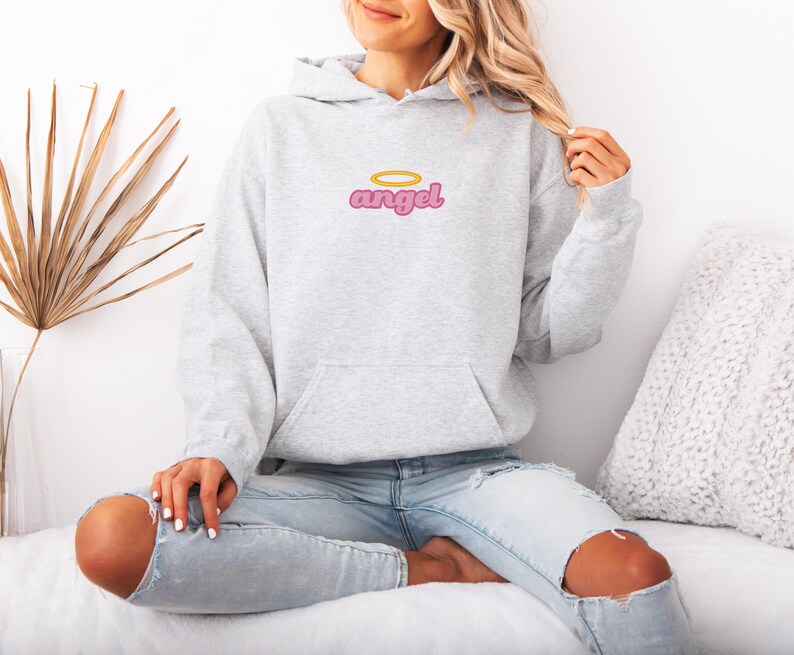 May include: A light gray hoodie with a front pocket and a hood. The word "angel" is embroidered on the chest in pink with a gold halo above it. The model is wearing light blue jeans with ripped knees.