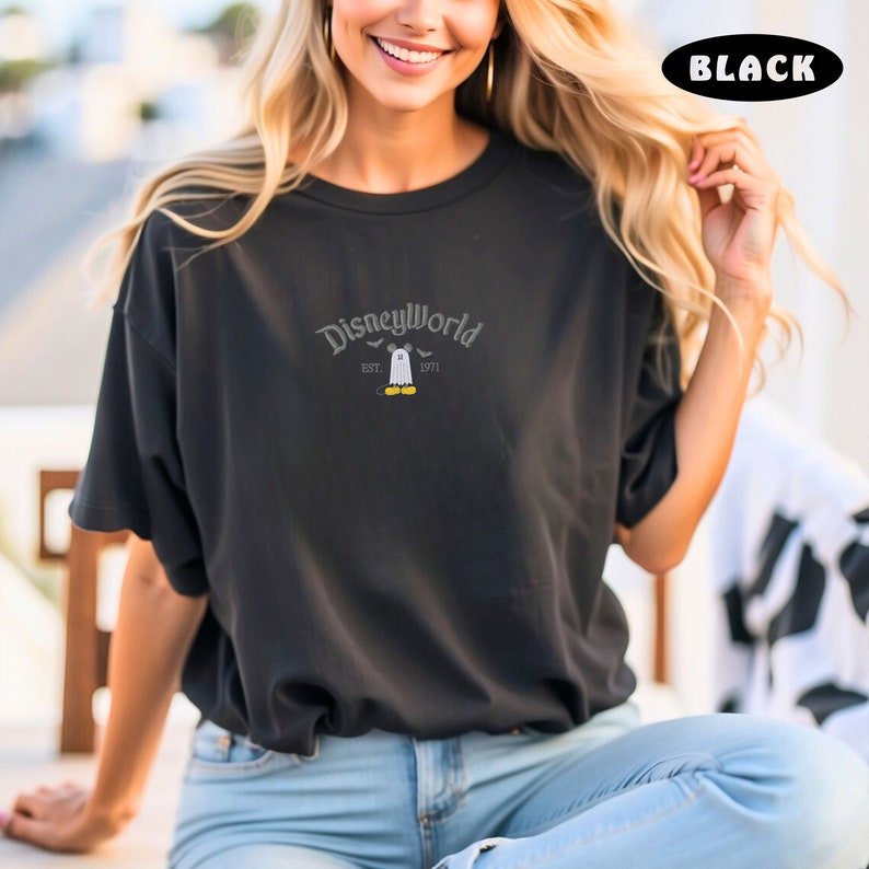 May include: Black short-sleeved t-shirt with the text "DisneyWorld" in a stylized font. The design includes a ghost and Mickey Mouse. The shirt is made of a soft material.