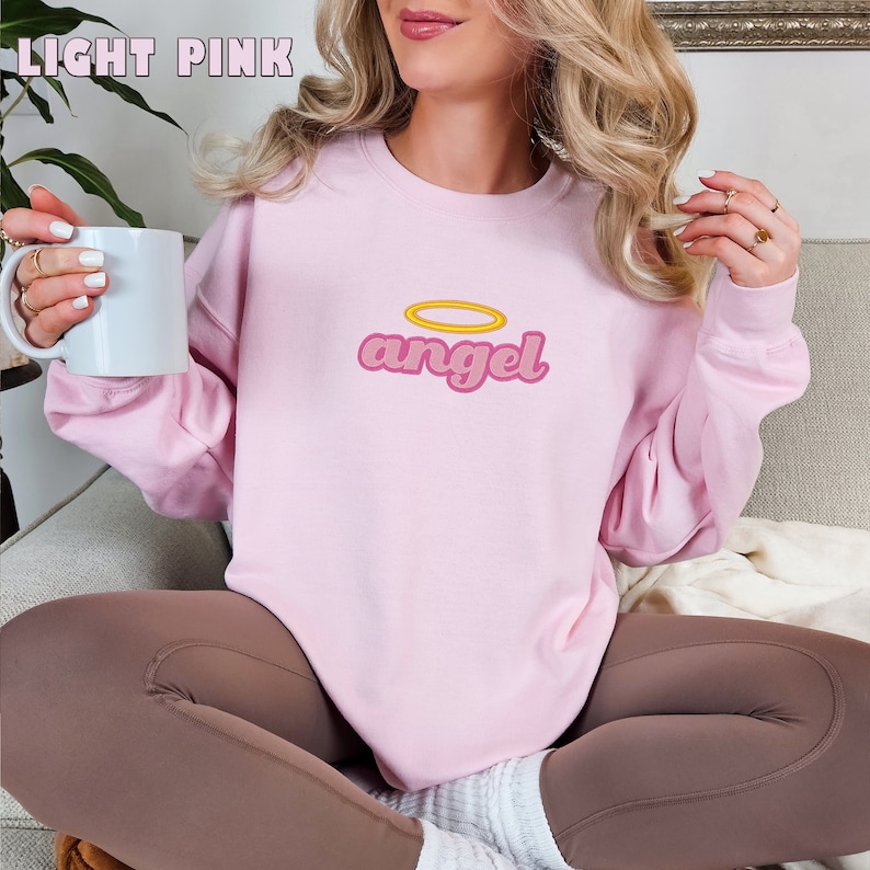 Puede incluir: Una sudadera de color rosa claro con un gr&aacute;fico retro de la palabra "angel" con un halo amarillo arriba.