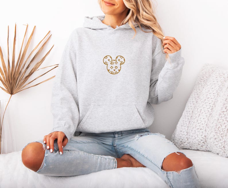 May include: A light gray hoodie with a front pocket and a drawstring hood. The hoodie features a gold-colored Mickey Mouse design on the chest. The design is filled with various shapes and patterns. The person is wearing light blue jeans.
