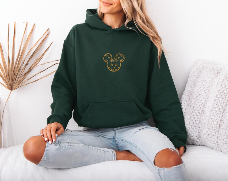 May include: A dark green hoodie with a front pocket and a hood. The hoodie features a gold-colored Mickey Mouse design on the chest. The design is made up of various shapes and patterns. The model is wearing light blue jeans.