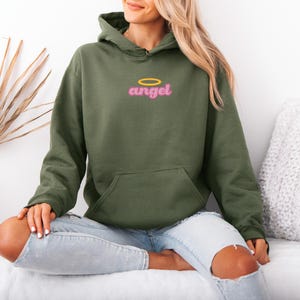 May include: An olive green hoodie with a front pocket and a hood. The word "angel" is printed on the chest in pink with a gold halo above it. The person is wearing light blue jeans.