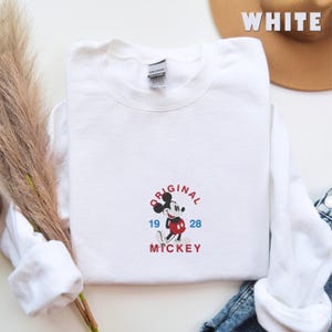 May include: A white crewneck sweatshirt featuring an embroidered graphic of Mickey Mouse in the center. The text reads "ORIGINAL" in red above the character, "19" in blue to the left, "28" in blue to the right, and "MICKEY" in red below.