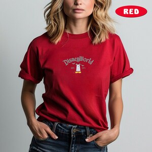 May include: A red t-shirt featuring the text "Disneyworld" above a cartoon ghost character with the text "EST. 1971". The shirt has rolled-up sleeves.