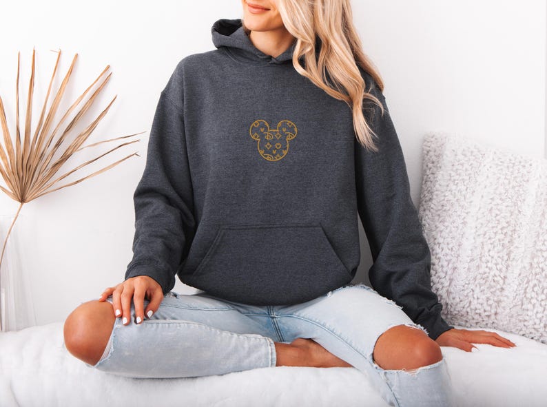 May include: Charcoal gray hoodie with a gold embroidered Mickey Mouse design on the front. The hoodie has a kangaroo pocket and a drawstring hood. The person is wearing light blue jeans. The background is white.