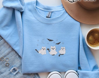 May include: Light blue sweatshirt with an embroidered design of three ghost cats, bats, a crescent moon, and stars. The text "Light Blue" is above the sweatshirt. The sweatshirt is folded on a white surface, next to a pair of gray sneakers, a cup of coffee, and a brown hat.