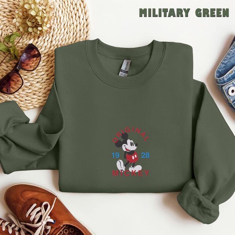 May include: A military green crewneck sweatshirt featuring an embroidered graphic of Mickey Mouse in the center. The text reads ORIGINAL MICKEY in red, with the numbers 19 and 28 in blue on either side of the character. The garment has long sleeves.