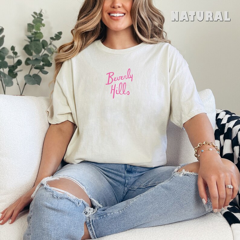 May include: A woman wearing a white t-shirt with the text "Beverly Hills" in pink lettering. She is sitting on a white couch wearing blue jeans.