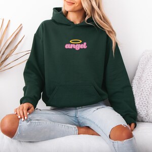 May include: Dark green hoodie with a front pocket and a hood. The word "angel" is embroidered in pink with a yellow halo above it. The hoodie is made of a soft material.