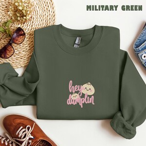 May include: A military green crewneck sweatshirt featuring an embroidered graphic of two smiling dumplings and the pink text "hey dumplin" centered on the chest. The garment has long sleeves, a ribbed neckline, and a standard fit construction.
