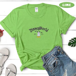 May include: Lime green t-shirt with rolled sleeves featuring the text "Disneyworld" in a dark gray font. Below the text is a graphic of Mickey Mouse dressed as a ghost, with the text "EST. 1971".