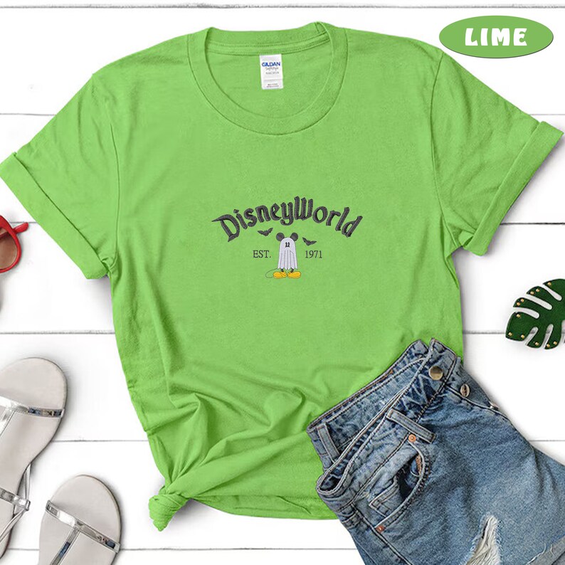 May include: Lime green t-shirt with rolled sleeves featuring the text "Disneyworld" in a dark gray font. Below the text is a graphic of Mickey Mouse dressed as a ghost, with the text "EST. 1971".
