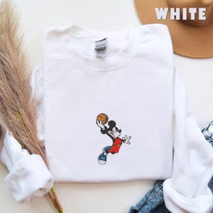 May include: A white crewneck sweatshirt featuring an embroidered graphic of a cartoon mouse wearing a red jersey and blue sneakers, mid-air while holding a basketball. The garment has a Gildan brand label at the inner neckline and long sleeves.