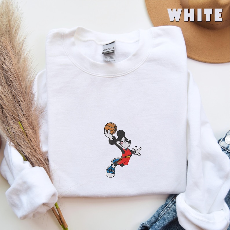 May include: A white crewneck sweatshirt featuring an embroidered graphic of a cartoon mouse wearing a red jersey and blue sneakers, mid-air while holding a basketball. The garment has a Gildan brand label at the inner neckline and long sleeves.