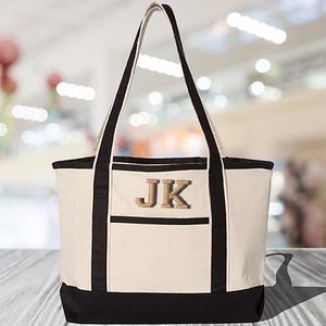 May include: A white canvas tote bag with black trim and a large pocket on the front. The bag has a black handle and the initials "JK" are embroidered on the pocket in a gold color.