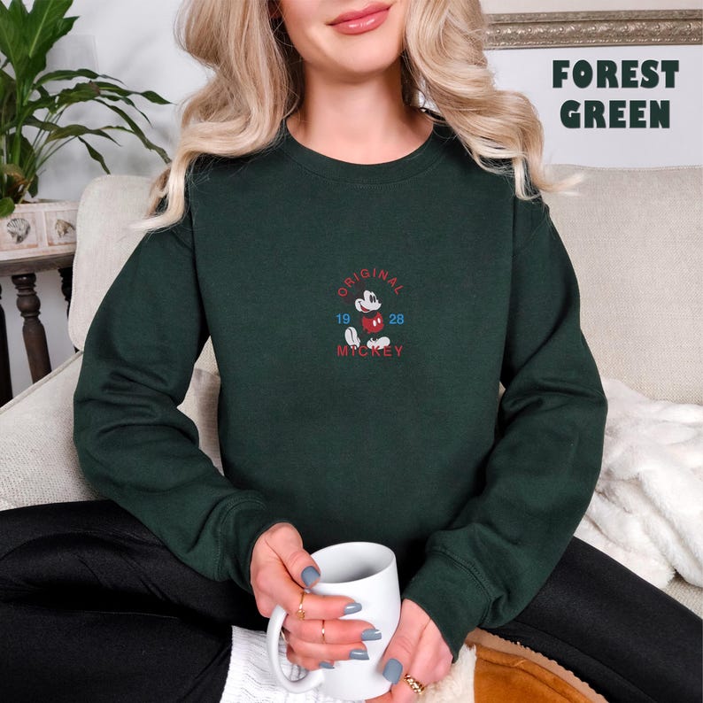 May include: A forest green long-sleeve sweatshirt featuring an embroidered graphic of Mickey Mouse in the center. The text reads ORIGINAL MICKEY 19 28 in red and blue thread. The sweatshirt has a crew neckline and ribbed cuffs and hem.