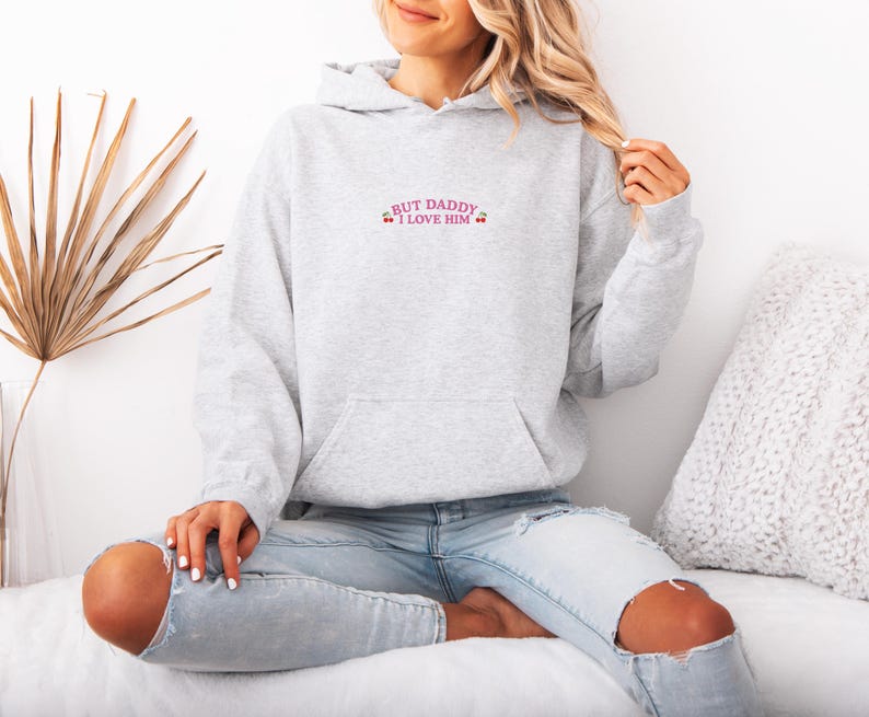 K&ouml;nnte beinhalten: Ein hellgrauer Kapuzenpullover mit einer Vordertasche und einer Kapuze. Der Text "BUT DADDY I LOVE HIM" ist in Rosa auf die obere Brust gestickt. Der Kapuzenpullover ist aus einem weichen Material.