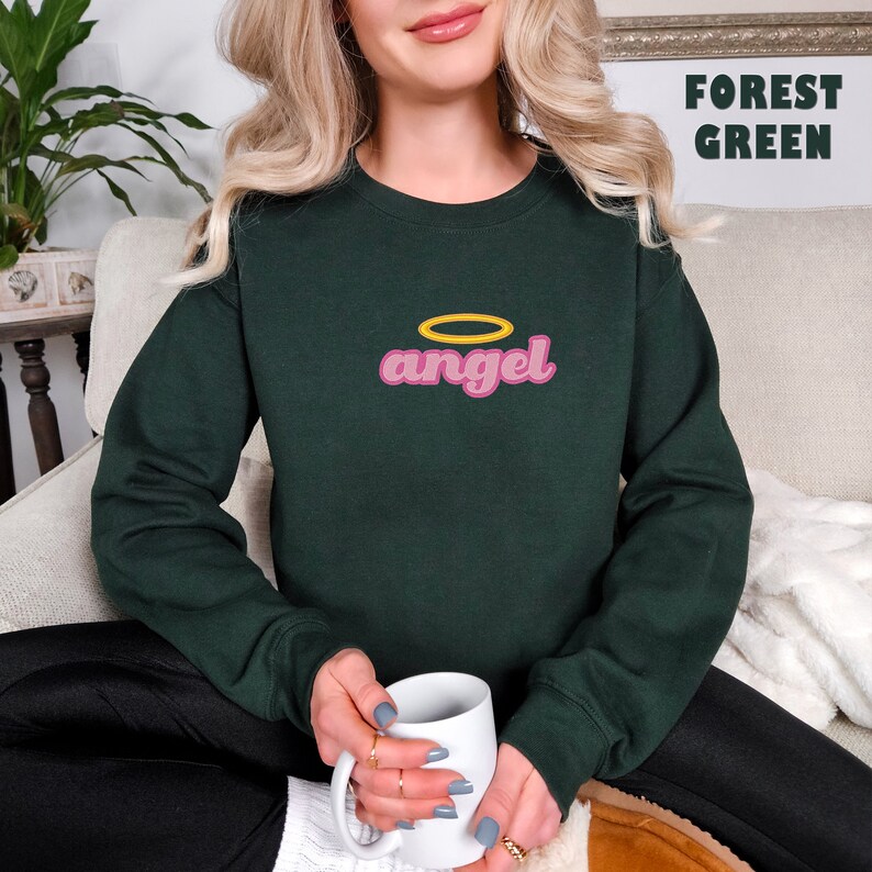 Puede incluir: Una sudadera verde bosque con la palabra "angel" en rosa y un halo amarillo arriba.