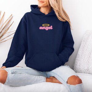 May include: Navy blue hooded sweatshirt with the word "angel" in pink script and a yellow halo above it. The sweatshirt has a front pocket and a drawstring hood. The model is wearing light blue jeans.