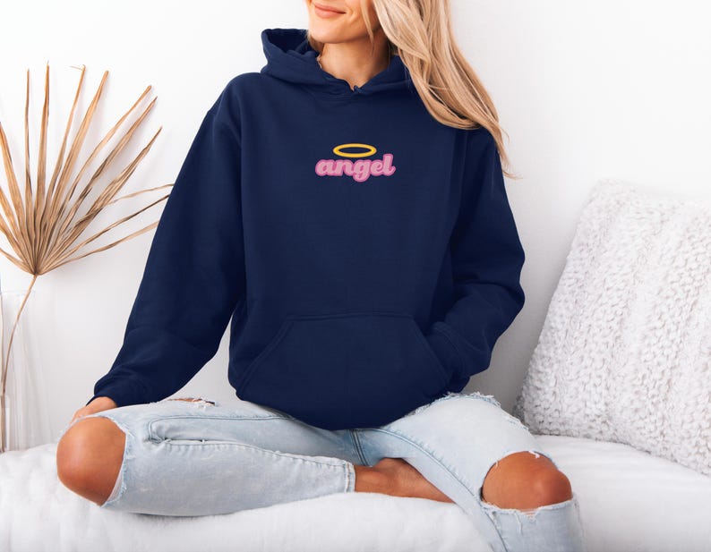 May include: Navy blue hooded sweatshirt with the word "angel" in pink script and a yellow halo above it. The sweatshirt has a front pocket and a drawstring hood. The model is wearing light blue jeans.