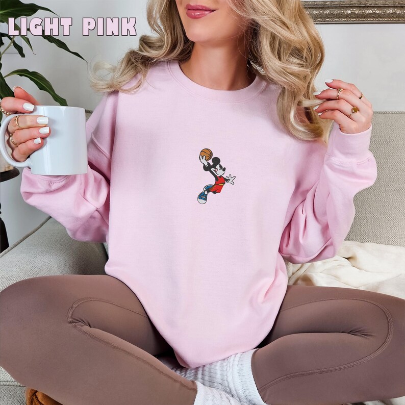 May include: A light pink long-sleeve crewneck sweatshirt featuring a small embroidered graphic of a cartoon mouse wearing a red jersey and blue sneakers while jumping to shoot a basketball. The text LIGHT PINK is printed in the upper left corner.