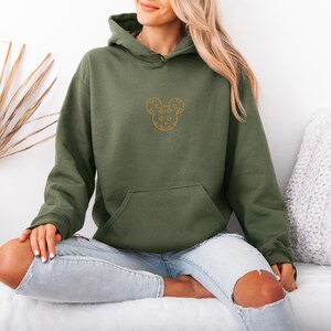 May include: Olive green hoodie with a kangaroo pocket and a drawstring hood. The hoodie features a gold-colored embroidered Mickey Mouse design on the chest. The model is wearing light blue jeans.