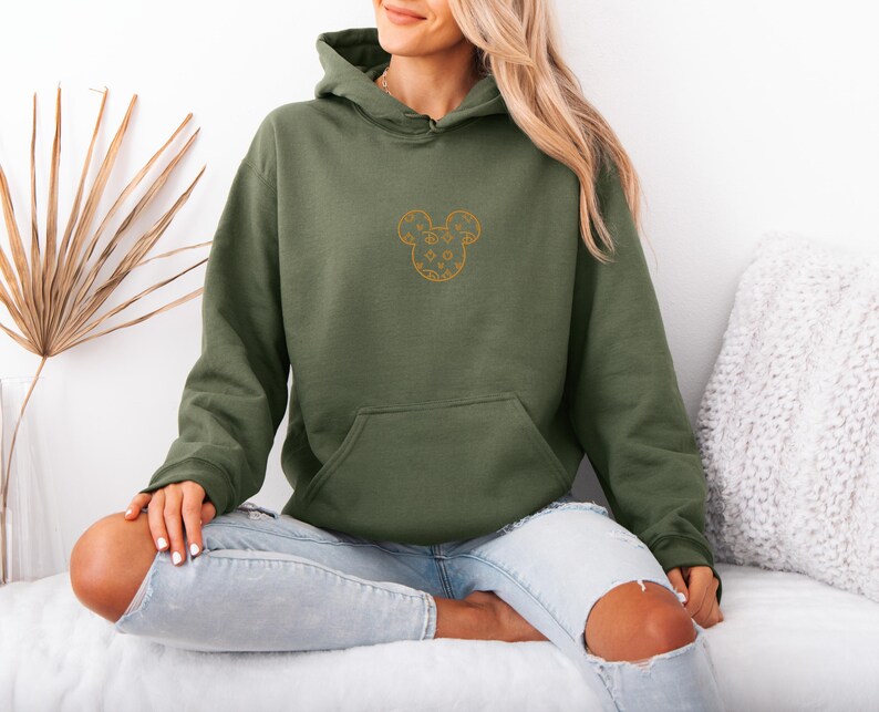 May include: Olive green hoodie with a kangaroo pocket and a drawstring hood. The hoodie features a gold-colored embroidered Mickey Mouse design on the chest. The model is wearing light blue jeans.
