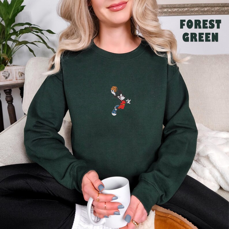 May include: A forest green crewneck sweatshirt featuring a small embroidered graphic of a cartoon character in a red jersey jumping to shoot a basketball. The text FOREST GREEN is printed in the upper right corner of the image.