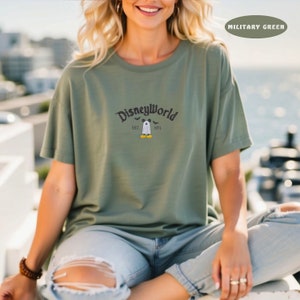 May include: Military green t-shirt with the text "DisneyWorld" in a curved font, with a ghost character and the text "EST. 1971". The shirt is a solid color and has short sleeves.