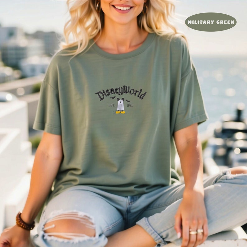May include: Military green t-shirt with the text "DisneyWorld" in a curved font, with a ghost character and the text "EST. 1971". The shirt is a solid color and has short sleeves.