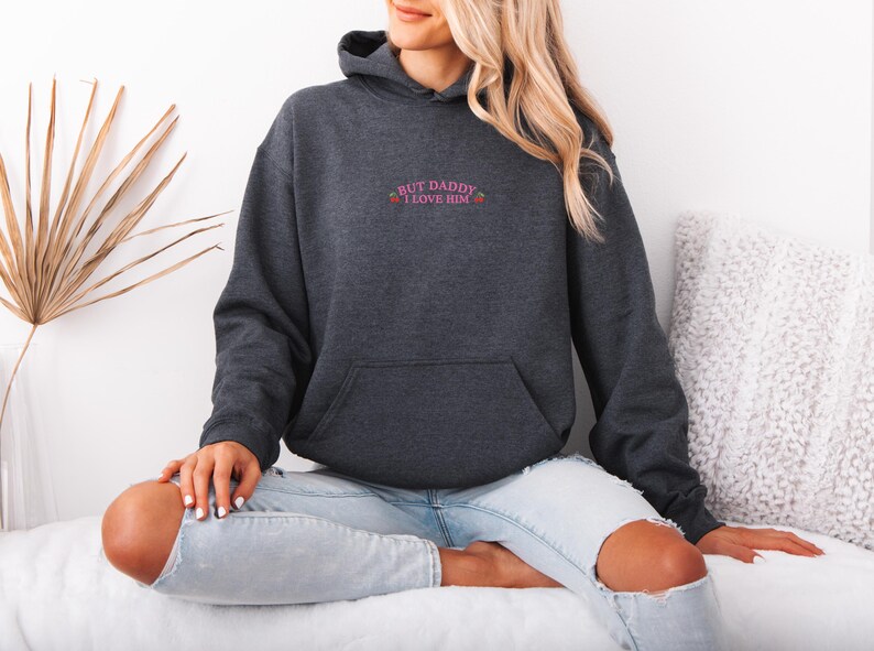 K&ouml;nnte beinhalten: Ein anthrazitfarbener Kapuzenpullover mit einer Vordertasche und einer Kapuze. Der Text "BUT DADDY I LOVE HIM" ist in Rosa &uuml;ber der Tasche gestickt. Das Model tr&auml;gt eine hellblaue Jeans.