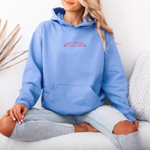 K&ouml;nnte beinhalten: Hellblauer Kapuzenpullover mit Fronttasche und Kapuze. Der Hoodie hat den gestickten Text "BUT DADDY I LOVE HIM" in Rosa. Das Model tr&auml;gt eine hellblaue, zerrissene Jeans.