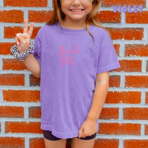 May include: A young person wearing a purple t-shirt with the text "Beverly Hills" in pink lettering. The person is standing in front of a brick wall.