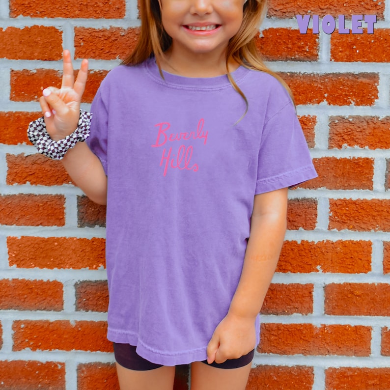 May include: A young person wearing a purple t-shirt with the text "Beverly Hills" in pink lettering. The person is standing in front of a brick wall.