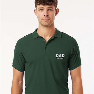 May include: Dark green polo shirt with the word "DAD - EST. 2025" printed in white on the chest. The shirt has a classic collar and short sleeves. The model is wearing a black belt.