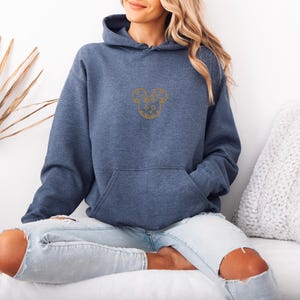 May include: A blue hooded sweatshirt with a kangaroo pocket and a gold embroidered design on the chest. The design is a stylized Mickey Mouse head. The sweatshirt is made of a soft material and is suitable for casual wear.