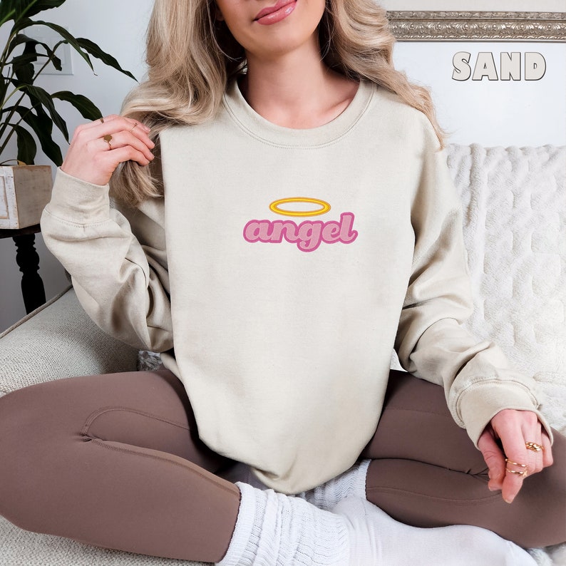Puede incluir: Una sudadera de color beige claro con un gr&aacute;fico de "angel" en rosa y un halo amarillo sobre el texto. La sudadera la lleva puesta una persona sentada en un sof&aacute;.