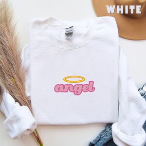 Puede incluir: Sudadera blanca con un gr&aacute;fico rosa de "angel" con un halo amarillo sobre la palabra.