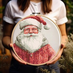 Puede incluir: Un aro de bordado de punto de cruz con una representación colorida de Santa Claus. El bordado presenta una barba blanca, un gorro rojo y una chaqueta con estampado rojo y blanco.