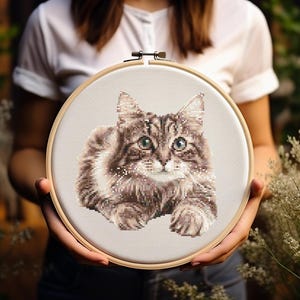 Könnte beinhalten: Kreuzstich-Stickerei einer Katze mit braun-weißem Fell, grünen Augen und einem flauschigen Aussehen. Die Katze ist auf einem beigefarbenen Stoff in einem hölzernen Stickrahmen zentriert. Das Kunstwerk wird hochgehalten.