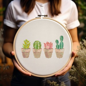 Può includere: Un cerchio da ricamo bianco con un disegno a punto croce di quattro piante grasse in vaso. Le piante grasse sono verdi, gialle, rosa e un cactus verde chiaro.